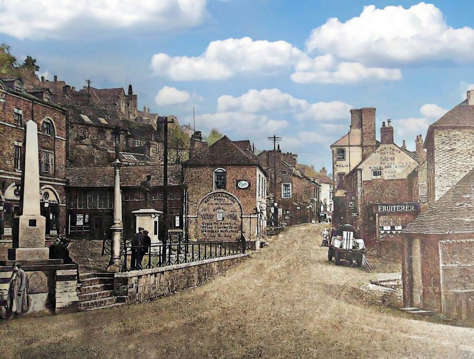 Looking across the Market Place towards the Tontine, the railed fountain on the left and a horse-drawn cart waiting on the setts. The road surface is the giveaway: no kerbs, no markings, just open ground shared by carts, stalls and pedestrians alike. Colourised; exact date unknown, but firmly the horse age.