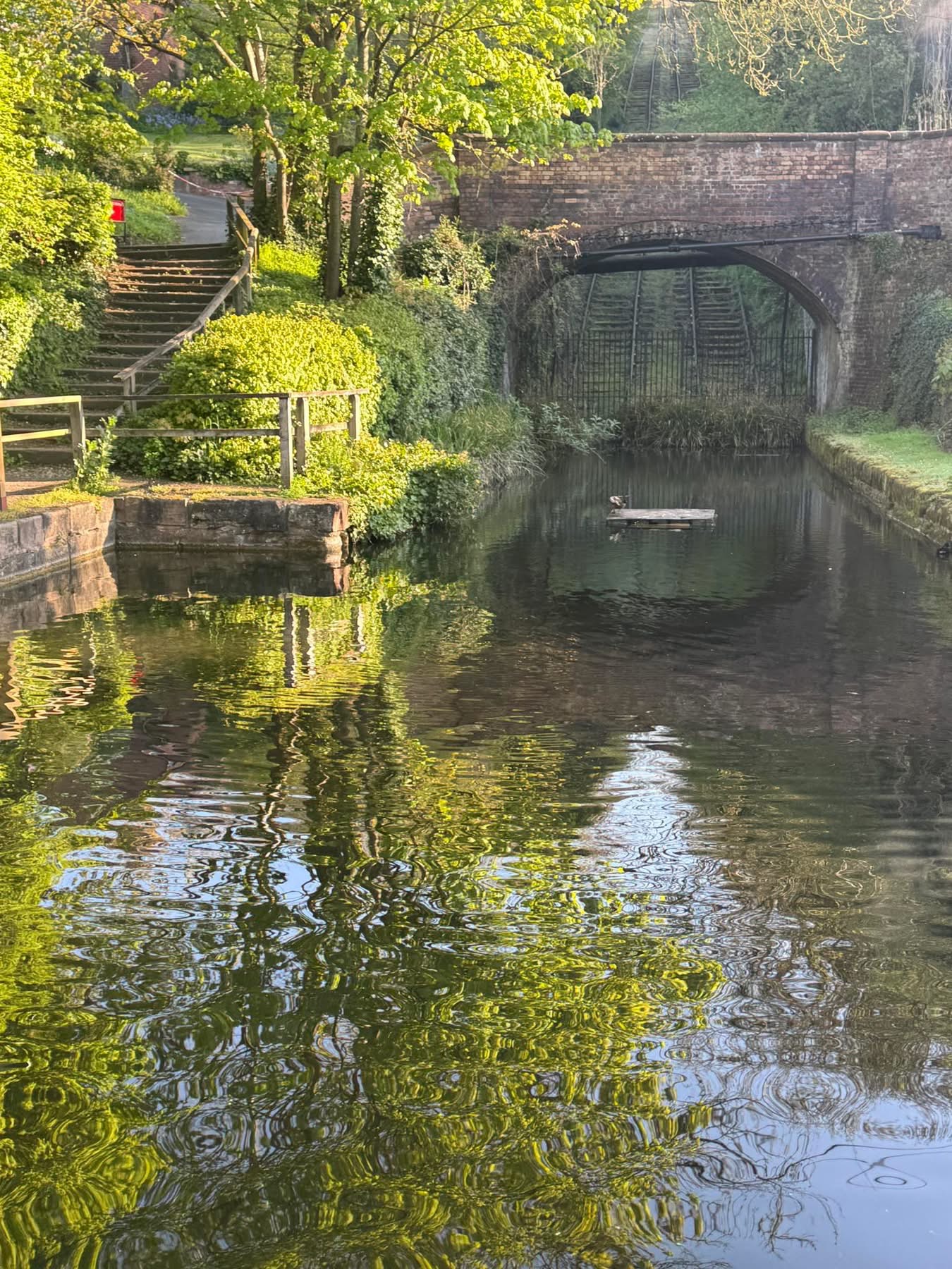 The canal basin at the head of the incline today, looking towards the Coalport High Street bridge, with the twin tracks climbing away beyond it. The basin holds water again, with steps and handrails now leading down to the top of the incline — a small, deliberate touch that lets present-day visitors stand exactly where the tub boats once floated.