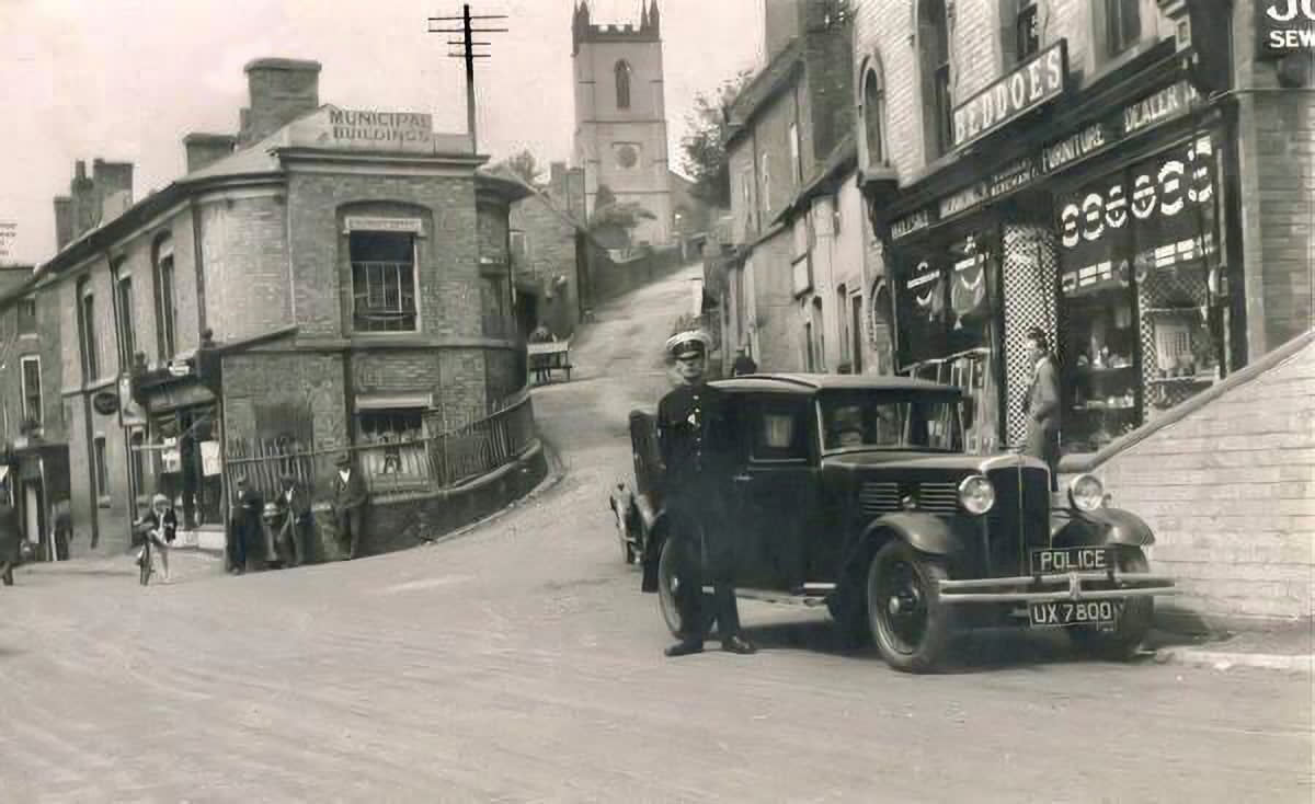 Historic street photograph with a police car parked in front of the Beddoes shop.