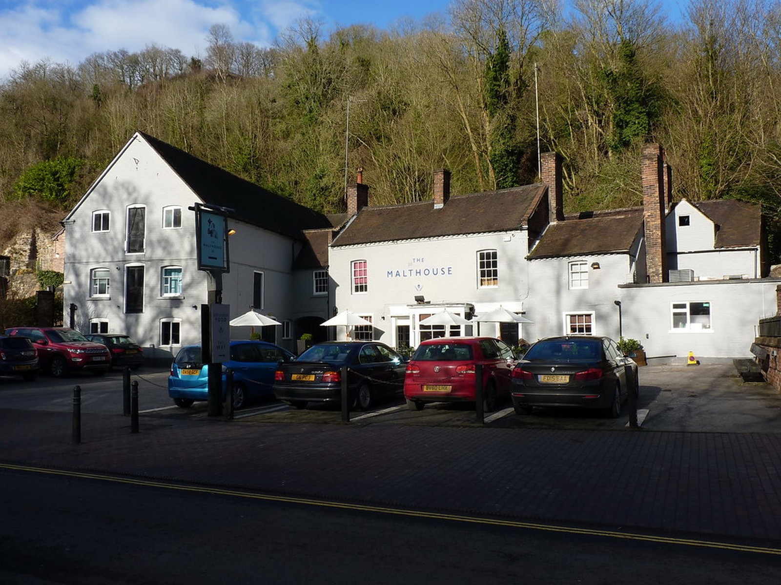 The Malthouse, Ironbridge, today: white-painted buildings on The Wharfage with parasols outside and wooded hillside behind